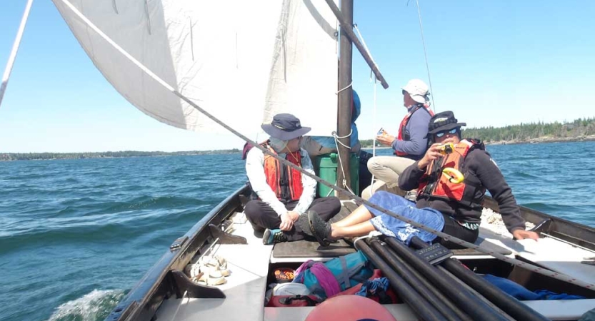 A group of students wearing life jacket sits in a sailboat on very blue water